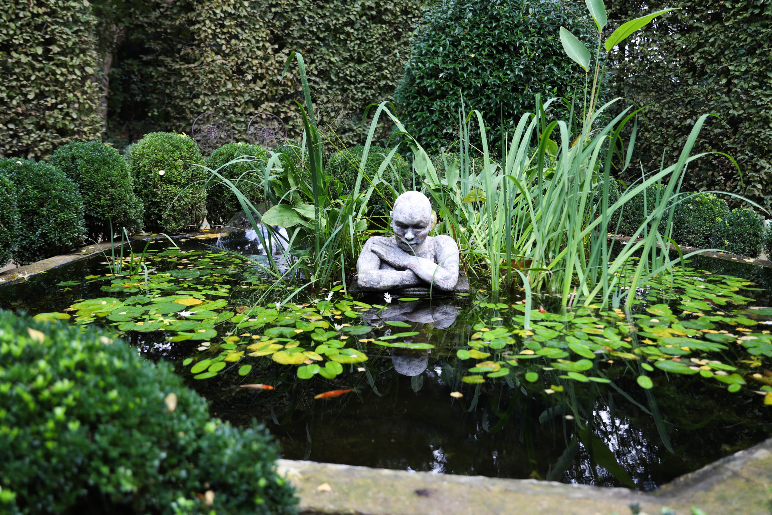 bassin avec, au milieu, une sculpture d'homme qui a les bras croisés et des plantes tout autour