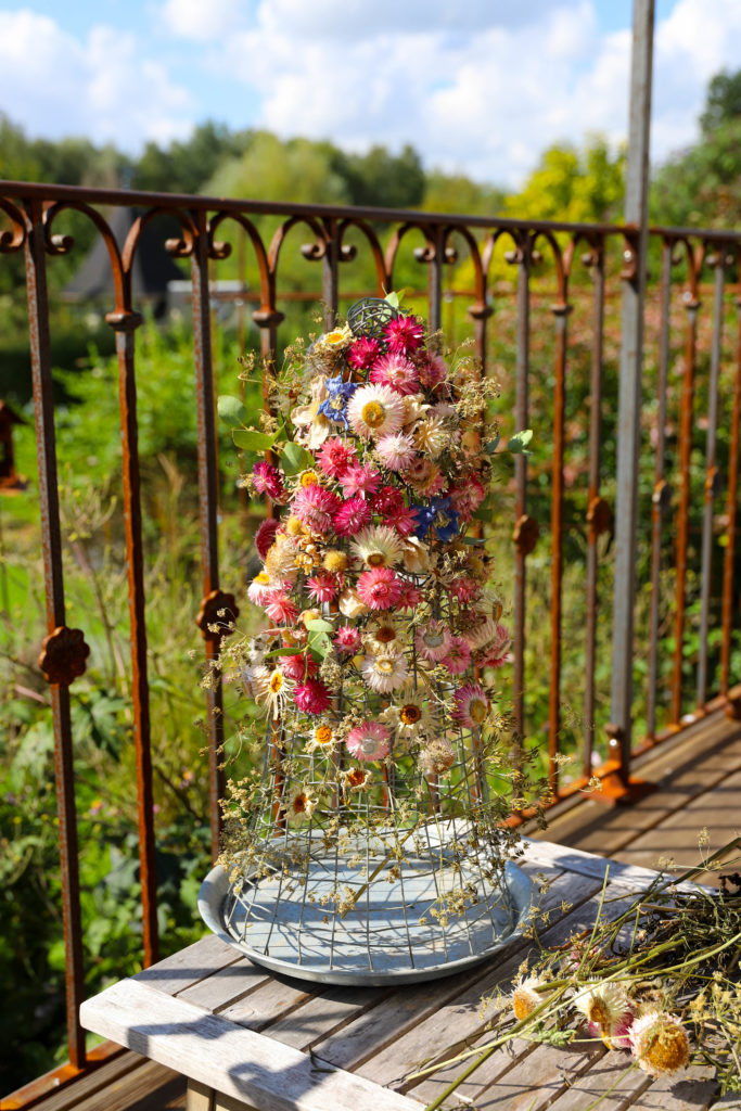 composition de fleurs séchées accrochées sur une cloche réalisée avec du grillage. La composition est posée sur une table en bois, derrière il y a une charmante balustrade en fer rouillé