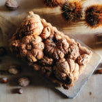 gâteau aux châtaignes qui a été réalisé dans un moule en métal avec une forme de fruits d'automne : pommes de pin, citrouilles. Le gâteau est doré. Il est posé sur une table en bois entouré de noix et de coques de châtaignes